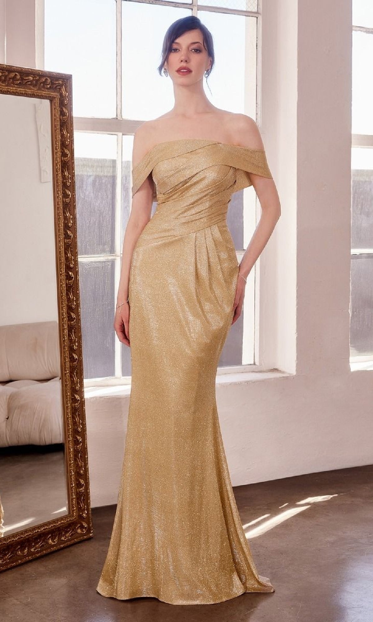 A woman stands indoors wearing the Amoris T603 Off-Shoulder Long Glitter Prom Dress, a floor-length gold evening gown with subtle shimmer and a mermaid skirt. A large framed mirror and a sofa are in the background.
